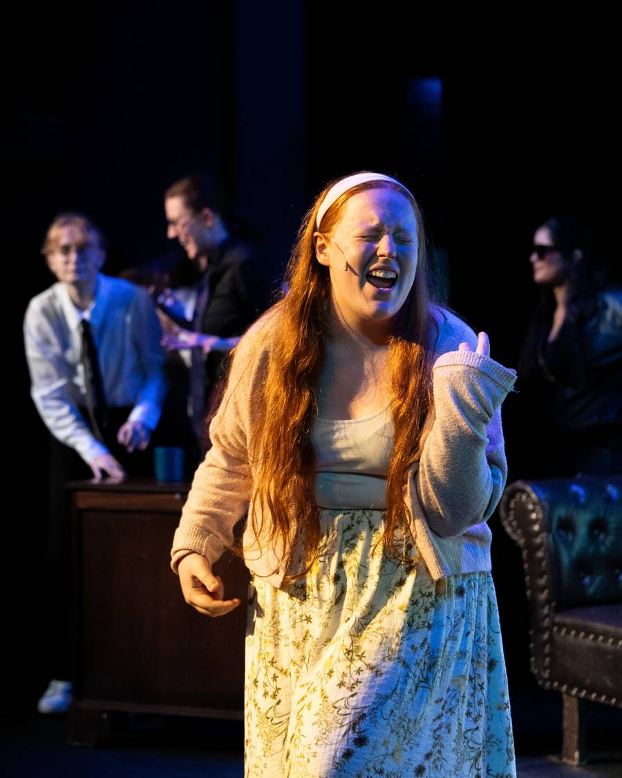 A performer in a yellow dress singing under stage lights with the cast behind her