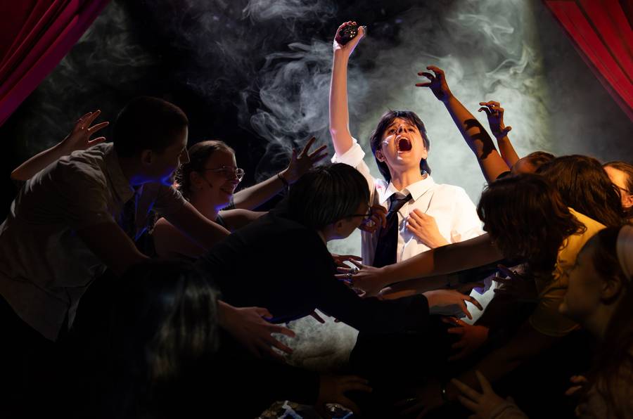 An ensemble reaching towards a performer in a white shirt amid stage smoke and red curtains